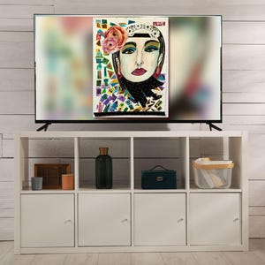 Puede incluir: Una colorida obra de arte de un rostro femenino se muestra en una gran pantalla de televisión. La obra presenta a una mujer con un tocado floral, labios rojos y una bufanda negra. El televisor está colocado sobre una unidad de almacenamiento blanca.