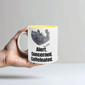 Op de afbeelding: Witte keramische mok met een gele binnenkant, met een cartoon kat illustratie en de tekst "Alert. Concerned. Caffeinated." De kat is grijs met zwarte accenten. De mok heeft een wit handvat.