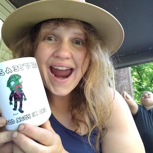 May include: White ceramic mug featuring a cartoon zombie character in a suit. The mug has the text "MOS A 33T" and "GOD AVOY 3N 130" printed on it. The person in the image is wearing a hat and smiling.