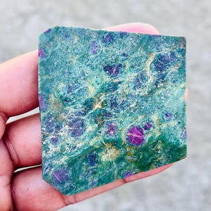 Könnte beinhalten: Ein rechteckiger, roher Edelstein mit einer türkis-grünen Matrix, übersät mit leuchtenden, tiefroten Rubinkristallen. Der Stein hat eine raue, natürliche Textur und unregelmäßige Kanten.