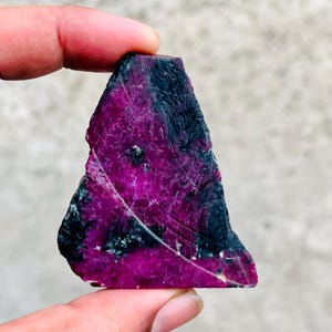 May include: A raw, triangular-shaped gemstone featuring a vibrant magenta and deep purple color palette with dark, opaque black inclusions and visible surface striations throughout the crystalline structure of the stone.