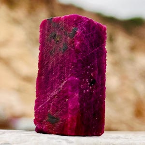 May include: A raw, rectangular ruby crystal with a deep magenta-pink hue. The stone features a rough, textured surface with visible natural striations and small, dark inclusions scattered across its vertical faces and top edge.