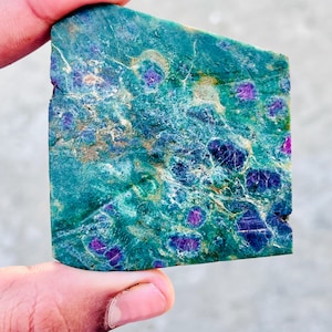 Op de afbeelding: Een hand houdt een rechthoekige steen vast met een turquoise en groene basiskleur. De steen heeft paarse en roze vlekken en aderen, met een ruw, getextureerd oppervlak. De steen heeft een natuurlijke, organische uitstraling.