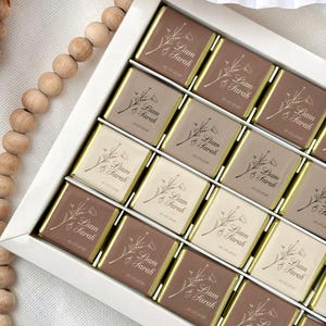 Pode incluir: Uma caixa branca cheia de chocolates embalados individualmente. Cada chocolate tem uma borda dourada e é impresso com "Liam & Sarah" e a data "10.05.2026", juntamente com um design floral. Os chocolates são em várias tonalidades de castanho e bege.