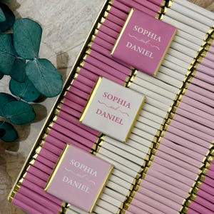 Op de afbeelding: Een verzameling kleine rechthoekige chocoladerepen verpakt in afwisselend roze en wit papier met goudfolie randen. Drie repen liggen bovenop, elk voorzien van de gedrukte tekst "SOPHIA and DANIEL" in een schreeflettertype.