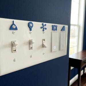 May include: A white light switch cover with blue decorative accents. The accents include a lightbulb, a fan, and other shapes. The light switches are in the on and off positions. The wall is painted a dark blue.