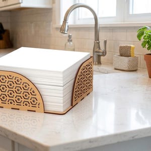 May include: A wooden paper holder with a geometric design holds a stack of white paper. The holder is made of light brown wood and sits on a white countertop in a kitchen setting. A small potted plant is visible in the background.