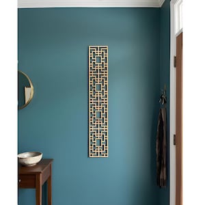 May include: A rectangular, light-colored wooden wall panel with a geometric openwork design. The panel is mounted on a teal wall, with a small wooden table and a decorative bowl in the foreground.