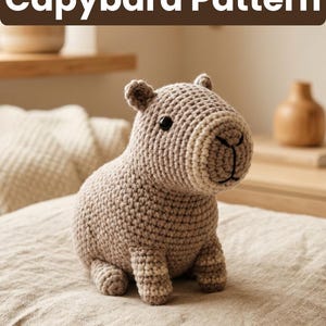 May include: A crocheted capybara toy in beige and brown tones is displayed. The toy features small ears, a black nose, and a stitched mouth. The text "Capybara Pattern" is above the toy. The toy is on a neutral-toned surface.