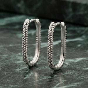 May include: A pair of silver oval hoop earrings. The earrings feature a textured, braided design along the outer edge. Each earring post is marked with "S925". The earrings are displayed on a dark green and grey marble surface.