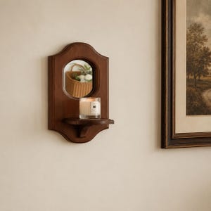 May include: A wall-mounted wooden mirror featuring a dark brown finish and a scalloped frame. The mirror includes a small, integrated shelf holding a clear glass candle jar with a white wax candle and a label.