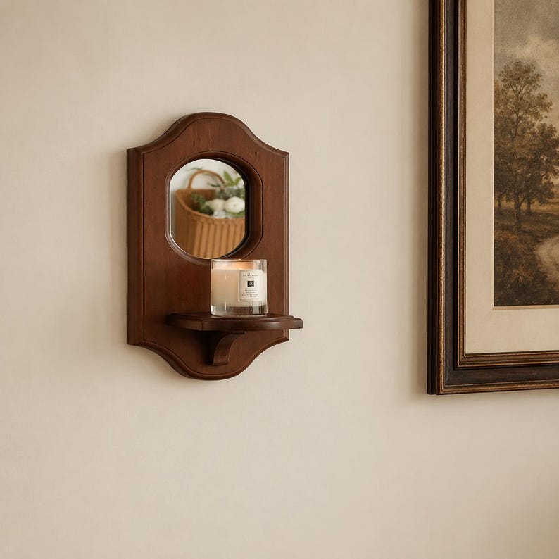 May include: A wall-mounted wooden mirror featuring a dark brown finish and a scalloped frame. The mirror includes a small, integrated shelf holding a clear glass candle jar with a white wax candle and a label.