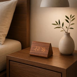 Puede incluir: Un reloj digital triangular de madera que muestra 22:18, sobre una mesita de noche de madera. A la derecha, un pequeño jarrón blanco con ramitas verdes. La escena está suavemente iluminada, creando un ambiente cálido.