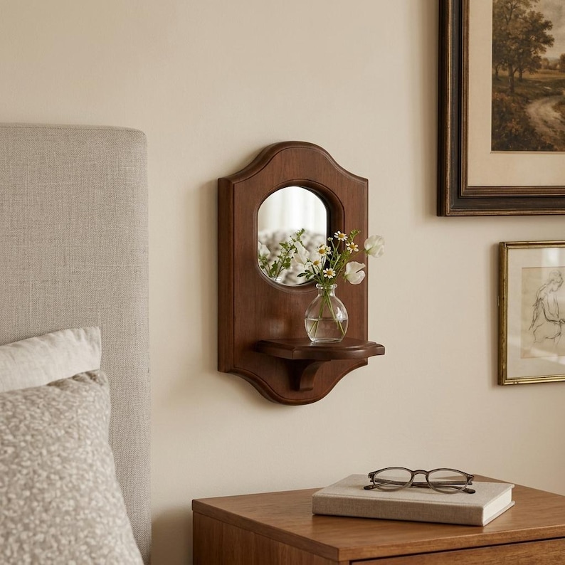 May include: A wall-mounted wooden mirror featuring a dark brown finish and a curved top edge. A small, clear glass vase containing white flowers sits on the integrated shelf below the circular mirror glass, reflecting the floral arrangement.