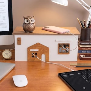 Op de afbeelding: Een wit en bruin houten oplaadstation in de vorm van een klein huisje met een plat dak. De voorzijde heeft een houten deur en raamuitsparing, terwijl op het bovenoppervlak een bronzen uilenbeeldje en een lichtroze smartphone zijn geplaatst.