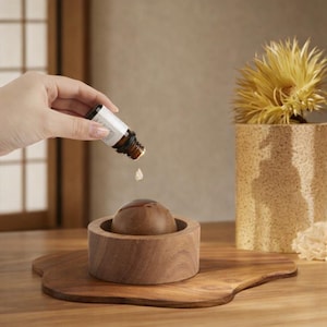 May include: A wooden essential oil diffuser with a round, brown wooden top and a circular base. A small bottle is pouring oil into the diffuser. The diffuser sits on a wooden tray, with a decorative plant in the background.