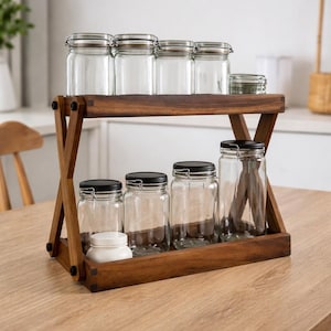 Puede incluir: Un especiero de madera de dos niveles con frascos de vidrio transparente y tapas negras. El estante contiene frascos de varios tamaños, perfectos para organizar especias de cocina. El estante de madera tiene un soporte en forma de X.
