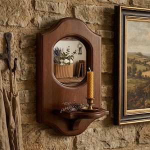 May include: A wall-mounted wooden mirror featuring a rounded top and a small integrated shelf. A tall, textured yellow beeswax candle sits in a brass holder on the shelf, accompanied by a small sprig of dried lavender. The mirror reflects a basket.