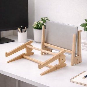 May include: A light-coloured wooden laptop stand with a minimalist design. The stand is angled to elevate a laptop, and is made of interlocking wooden pieces. The stand is on a white surface.