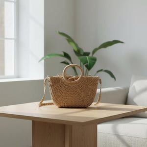 May include: A woven straw handbag with a circular handle and a shoulder strap sits on a wooden table. The bag is a light tan color and has a rounded shape. A plant with large green leaves is in the background.