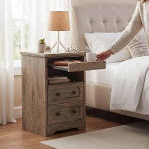 May include: A wooden nightstand with two drawers and a pull-out tray. The nightstand has a light brown finish and black knobs. A lamp, plant, and glasses sit on top. A person is reaching for a cup on the tray.