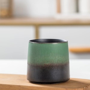 May include: A ceramic tumbler with a gradient design. The top half is a muted green, transitioning to a dark brown band, and ending in a black base. The interior rim is black. The tumbler sits on a wooden surface.