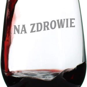 May include: A stemless wine glass being filled with red wine. The words "NA ZDROWIE" are printed on the glass. The wine is a deep red color, and the glass is clear.