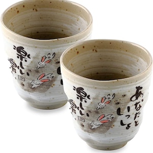 May include: Two ceramic cups with a beige and white speckled finish. Each cup features black Japanese characters and cartoon rabbits with pink ears. The cups have a rustic, handmade appearance.
