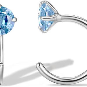 May include: A pair of silver-colored nose rings. Each ring features a light blue, round gemstone. One ring is shown from the side, displaying the curved post and the gemstone's setting. The other ring is shown from the front.
