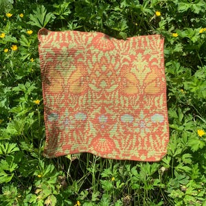 Puede incluir: Un cojín cuadrado tejido a ganchillo con un patrón floral repetido en tonos naranja, amarillo y verde. El cojín está sobre un lecho de follaje verde con pequeñas flores amarillas.