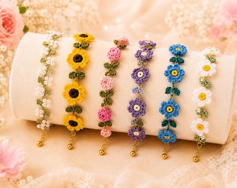 May include: Six handmade crochet bracelets featuring floral designs in white, yellow, pink, purple, and blue. Each bracelet displays distinct flower patterns with green leaves and a gold-toned metal chain extension ending in a small round bead.