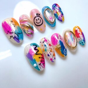 May include: A set of ten press-on nails featuring a variety of artistic designs, including vibrant multi-colored paint splatters, abstract brush strokes, gold metallic accents, a black smiley face, and gold-toned oval frames on a glossy finish.