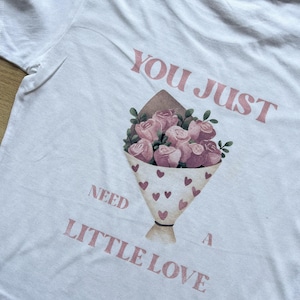 Peut inclure: T-shirt blanc avec un motif de bouquet floral. Le bouquet présente des roses roses et des feuilles vertes, enveloppé dans un cône en papier orné de cœurs. Le texte sur le t-shirt dit "YOU JUST NEED A LITTLE LOVE."