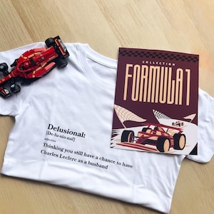 Op de afbeelding: Wit T-shirt met de definitie van "Delusional" erop gedrukt, samen met de tekst "Thinking you still have a chance to have Charles Leclerc as a husband." Een rode speelgoed raceauto en een Formula 1 Collection kaart zijn ook zichtbaar.