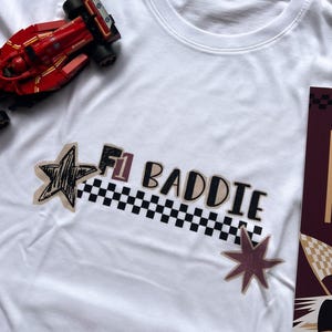 Peut inclure: T-shirt blanc avec l'inscription "F1 BADDIE" en noir et beige, avec une bande à carreaux noirs et blancs. Le t-shirt comporte des écussons en forme d'étoile et un jouet de voiture de course rouge.
