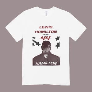 Peut inclure: T-shirt blanc avec un graphisme de Lewis Hamilton. Le design comprend le nom "LEWIS HAMILTON" en bordeaux, le numéro "44" et le mot "HAMILTON" en bas. Le t-shirt comporte également des graphismes d'étoiles.