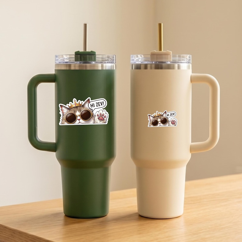 May include: Two insulated tumblers with handles. One is dark green, the other is beige. Each has a stainless steel lid, a straw, and a sticker of a cat wearing sunglasses and a crown, with the text "Hi Zev!"