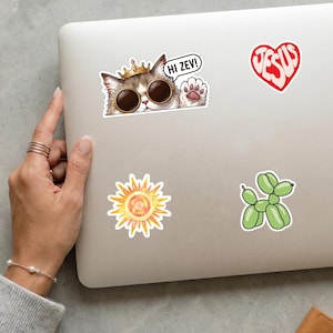 May include: A laptop adorned with stickers. One features a cat in sunglasses and a crown, with the text "HI ZEV!". Other stickers include a red heart with "JESUS" and a green balloon dog.