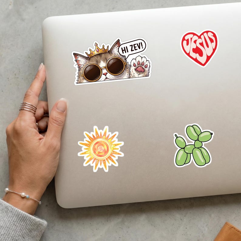May include: A laptop adorned with stickers. One features a cat in sunglasses and a crown, with the text "HI ZEV!". Other stickers include a red heart with "JESUS" and a green balloon dog.