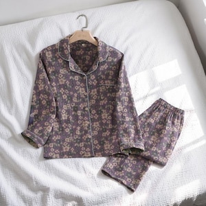 May include: A two-piece pajama set featuring a floral pattern in shades of purple, beige, and green. The long-sleeved top has a button-down front and white trim. The matching pants have an elastic waistband. The set is displayed on a white bed.
