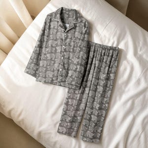 May include: Grey pyjama set featuring a repeating white bear design. The long-sleeved top has a button-down front and white trim. The matching trousers have an elasticated waistband. The set is displayed on a white bed.