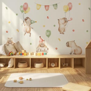 Könnte beinhalten: Ein Set Wandaufkleber mit Aquarellillustrationen von Wasserschweinen mit Luftballons, Partyhüten und Wimpelketten. Ein Wasserschwein hält ein Schild mit dem Namen Charlie. Die Aufkleber sind an einer Wand über einem Holzregal angebracht.