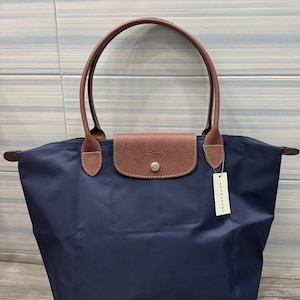 Puede incluir: Bolso tote azul marino con asas de cuero marrón y cierre de solapa. El bolso presenta un pequeño botón dorado en la solapa y una etiqueta que dice "LONGCHAMP". El bolso está hecho de un material duradero y resistente al agua.