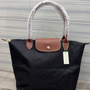 Op de afbeelding: Zwarte tote bag met bruine lederen accenten, waaronder een klepsluiting en handvatbevestigingen. De tas heeft een lange, gebogen handgreep en een klein label met "LONGCHAMP".