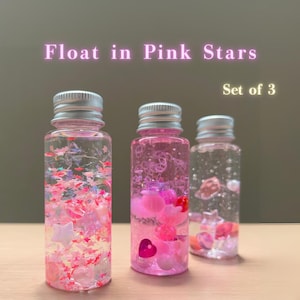 May include: Three clear bottles with silver lids, filled with pink liquid and floating star and heart-shaped decorations. The text "Float in Pink Stars" and "Set of 3" are visible.