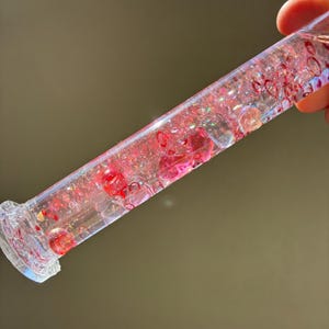 May include: A clear, cylindrical sensory toy filled with pink and red beads, glitter, and heart-shaped confetti. The toy has a decorative, textured base and is held at an angle, catching the light.