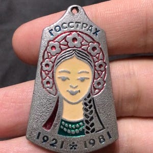 May include: A silver-toned metal pendant featuring a textured surface and a central enamel illustration of a person wearing a floral headpiece. The text "ГОССТРАХ" is at the top, with "1921" and "1981" at the bottom, separated by a small star symbol.
