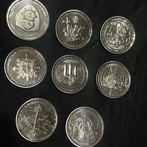 May include: A collection of nine silver coins, each with detailed relief designs. The coins feature various emblems, figures, and text, likely commemorative or collectible items. The coins are arranged on a dark surface.