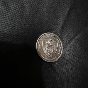 May include: A silver coin with embossed text and a central emblem featuring a flame and a starburst design. The coin's edge has additional text. The coin is set against a dark, textured background.