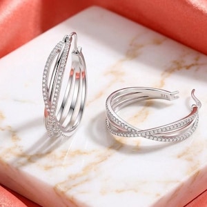 May include: A pair of silver hoop earrings featuring a triple-band crossover design. Each earring is accented with small, clear pavé-set stones along the front bands. The metal is stamped with 925, indicating sterling silver composition.